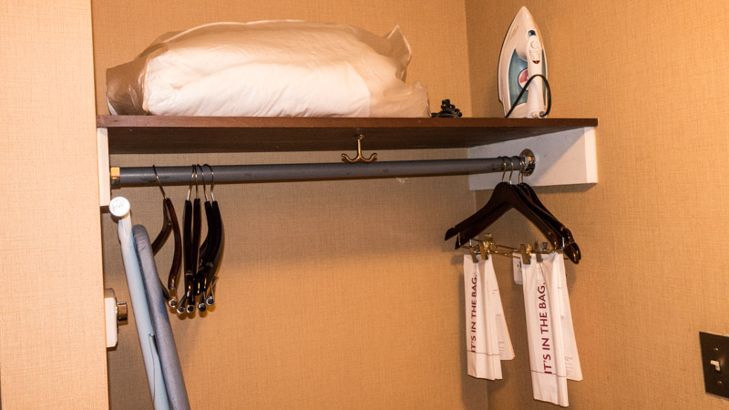Open closet with clothes hangers, iron, ironing board, extra blanket, and luggage rack